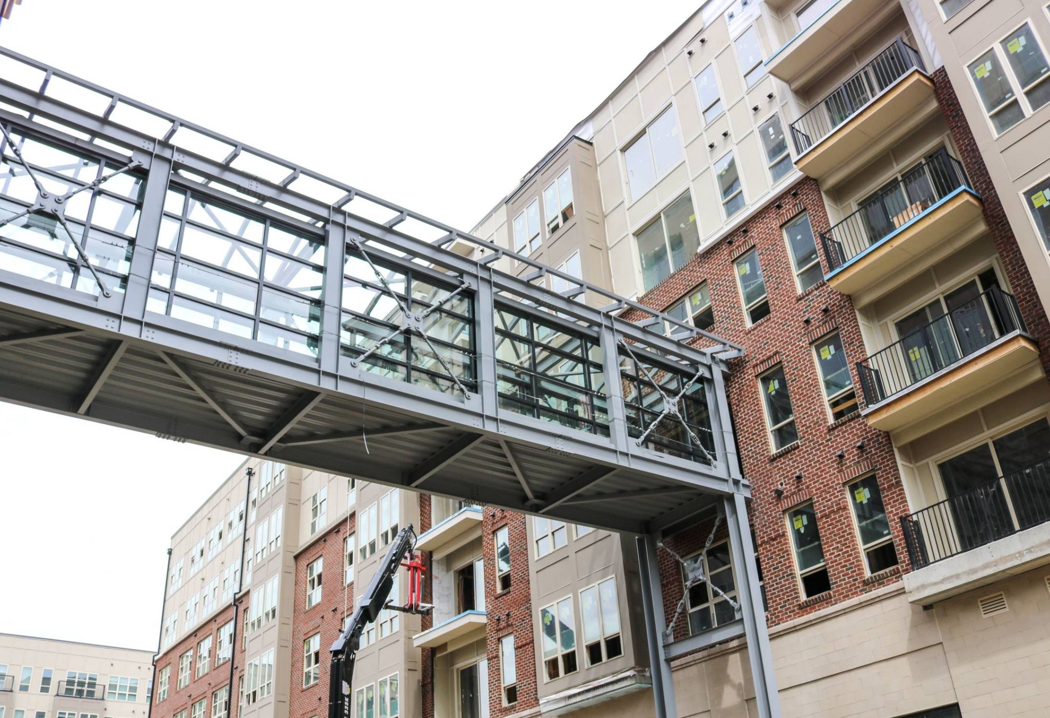 Glass skywalk connecting two modern apartment buildings with balconies, seen from below on a cloudy day.