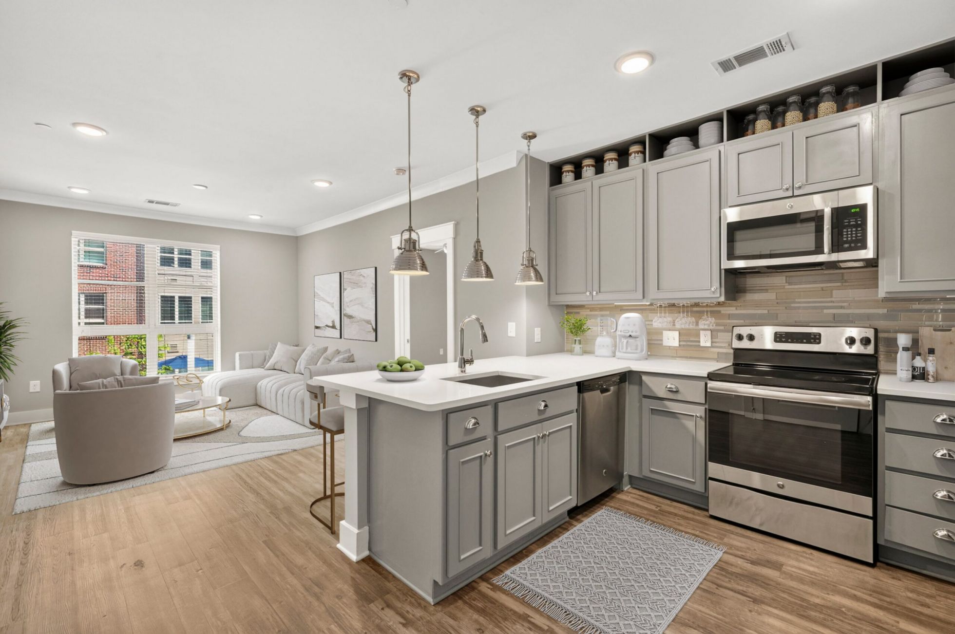 Modern kitchen with gray cabinets, stainless appliances, white countertops, pendant lights, and an open view to a bright living room.