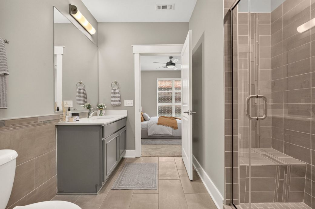 Modern bathroom with a glass shower, gray vanity, large mirror, and view into a bedroom with a ceiling fan and window.