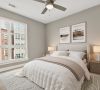 Modern bedroom with beige walls, large window, bed with white bedding, nightstands, lamps, art above headboard, and ceiling fan.