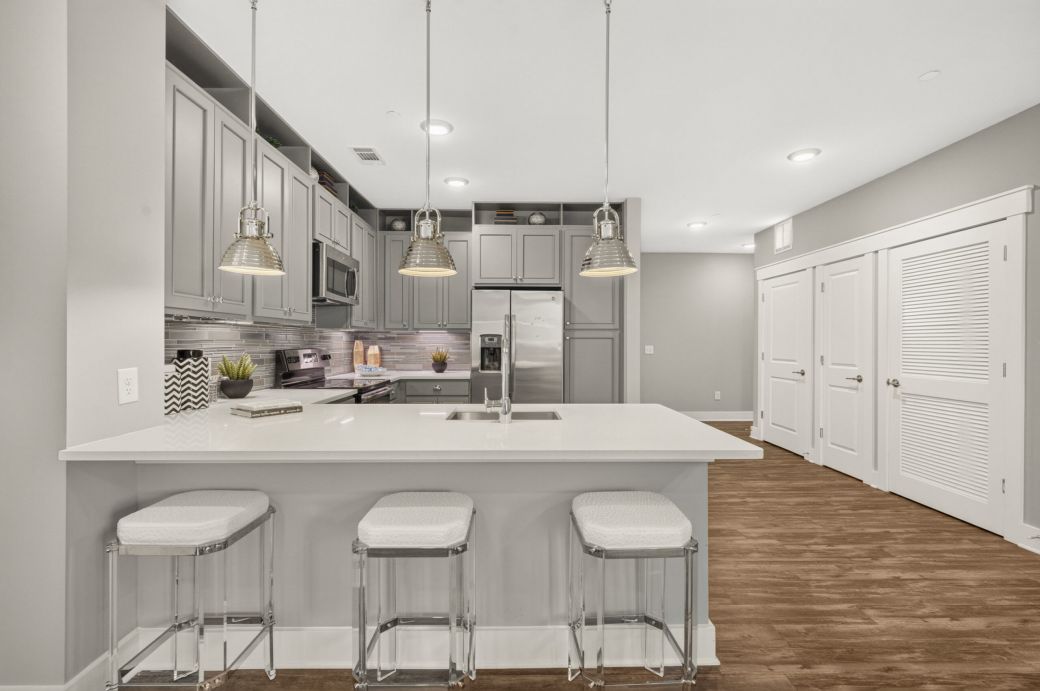 Modern kitchen with gray cabinets, stainless steel appliances, island with sink, and three clear stools on wood flooring.