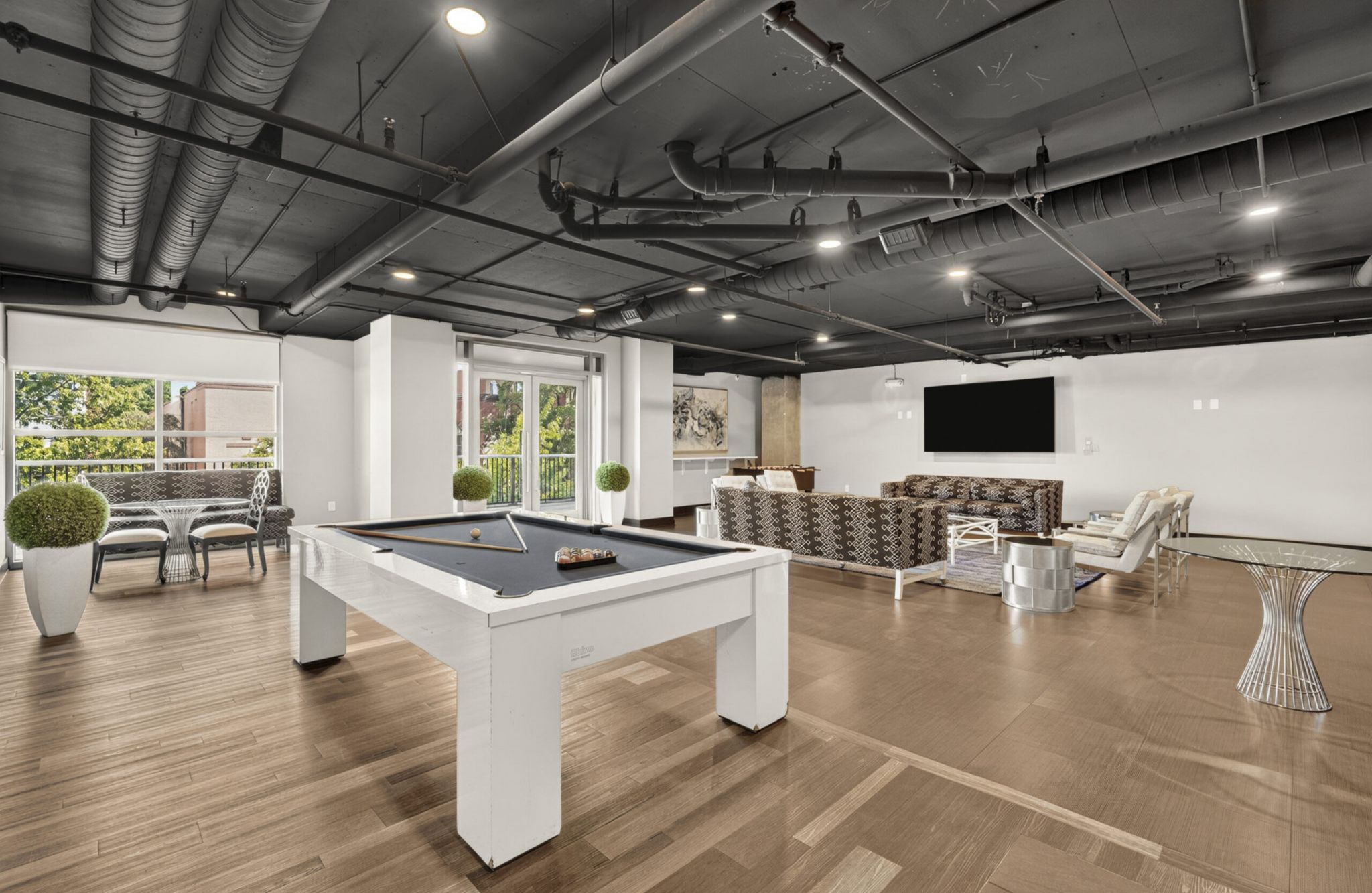 Modern lounge with a white pool table, seating area, large TV, and exposed ceiling pipes, plus windows showing greenery outside.