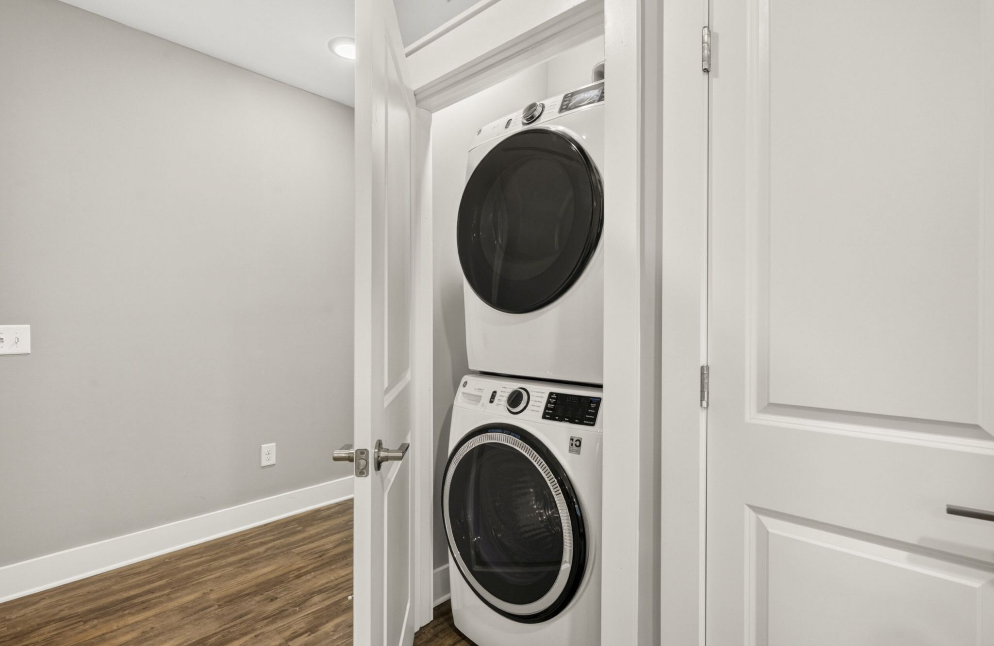A stacked washer and dryer set is inside a small closet with white doors in a laundry area with wood flooring.