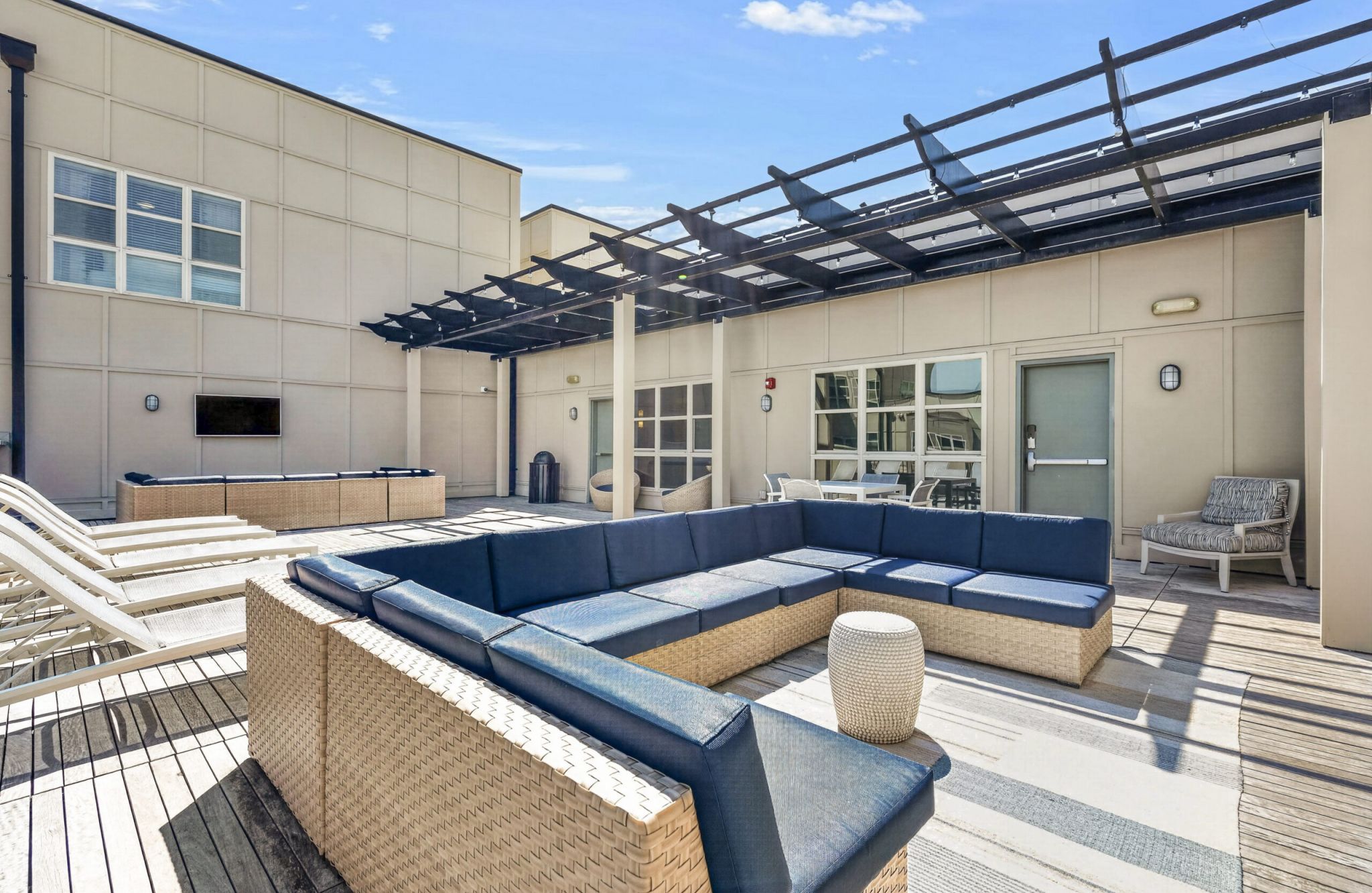Outdoor patio with wicker sectional sofas, blue cushions, lounge chairs, and a pergola on a wooden deck under a sunny sky.