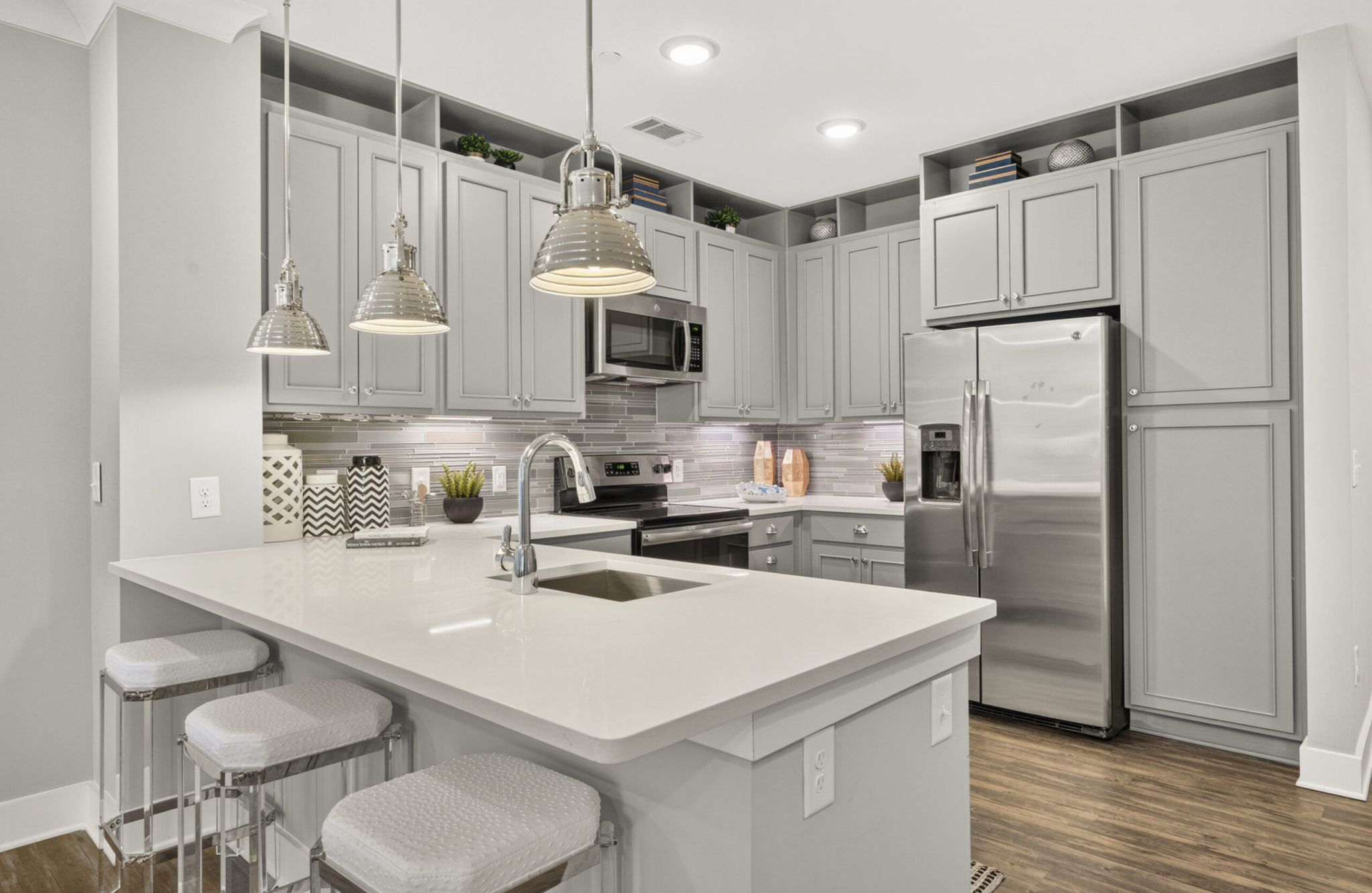 Modern kitchen with gray cabinets, stainless steel appliances, island with sink, pendant lights, and three stools on a wooden floor.